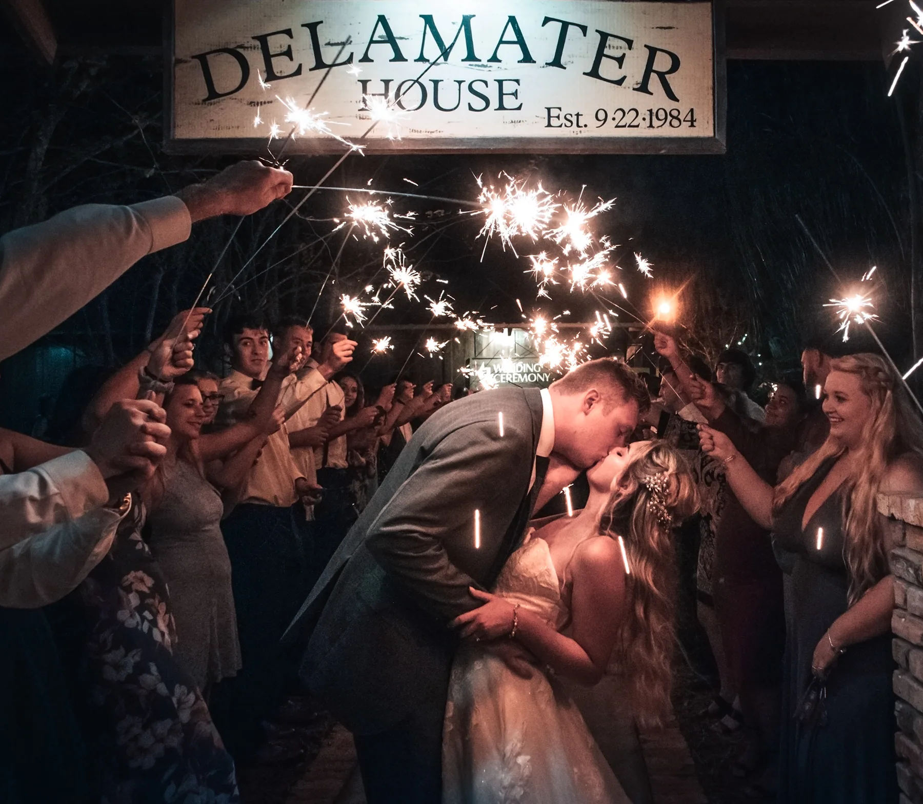 Newlyweds kissing during a sparkler exit beneath The Delamater House sign